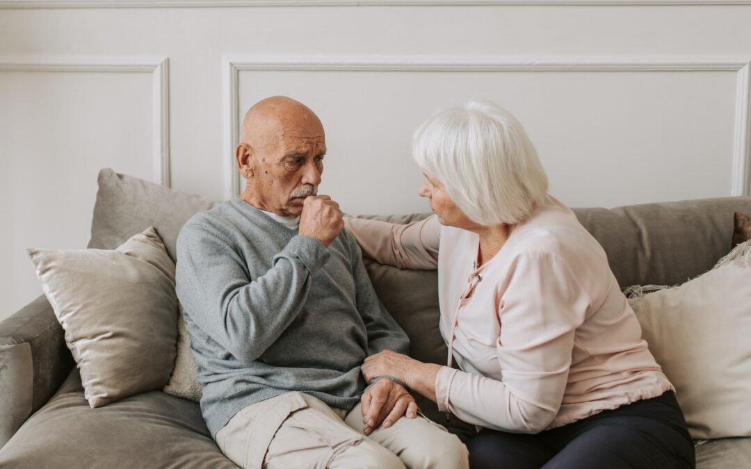 Elderly male coughing while a concerned female sitting with him watches on.