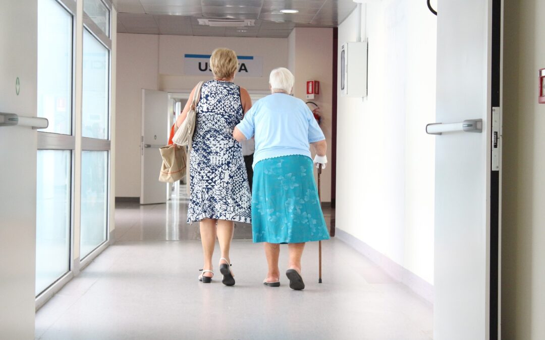 A back shot of a senior with a cane walking down a hospital corridor while also being supported by a younger lady.