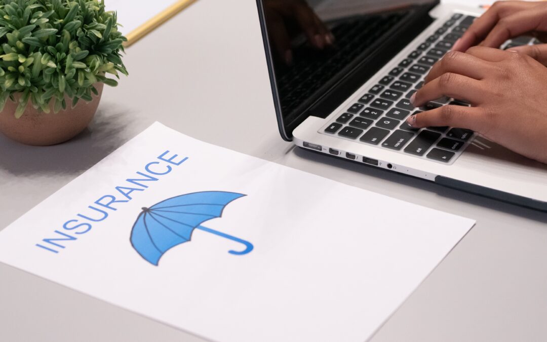 A person uses a laptop. There is a piece of paper with a blue umbrella and the word insurance above it.
