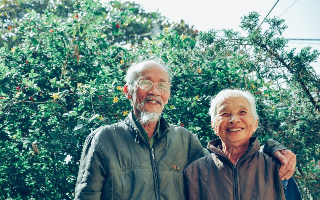 A smiling elderly couple is standing in front of a tree.