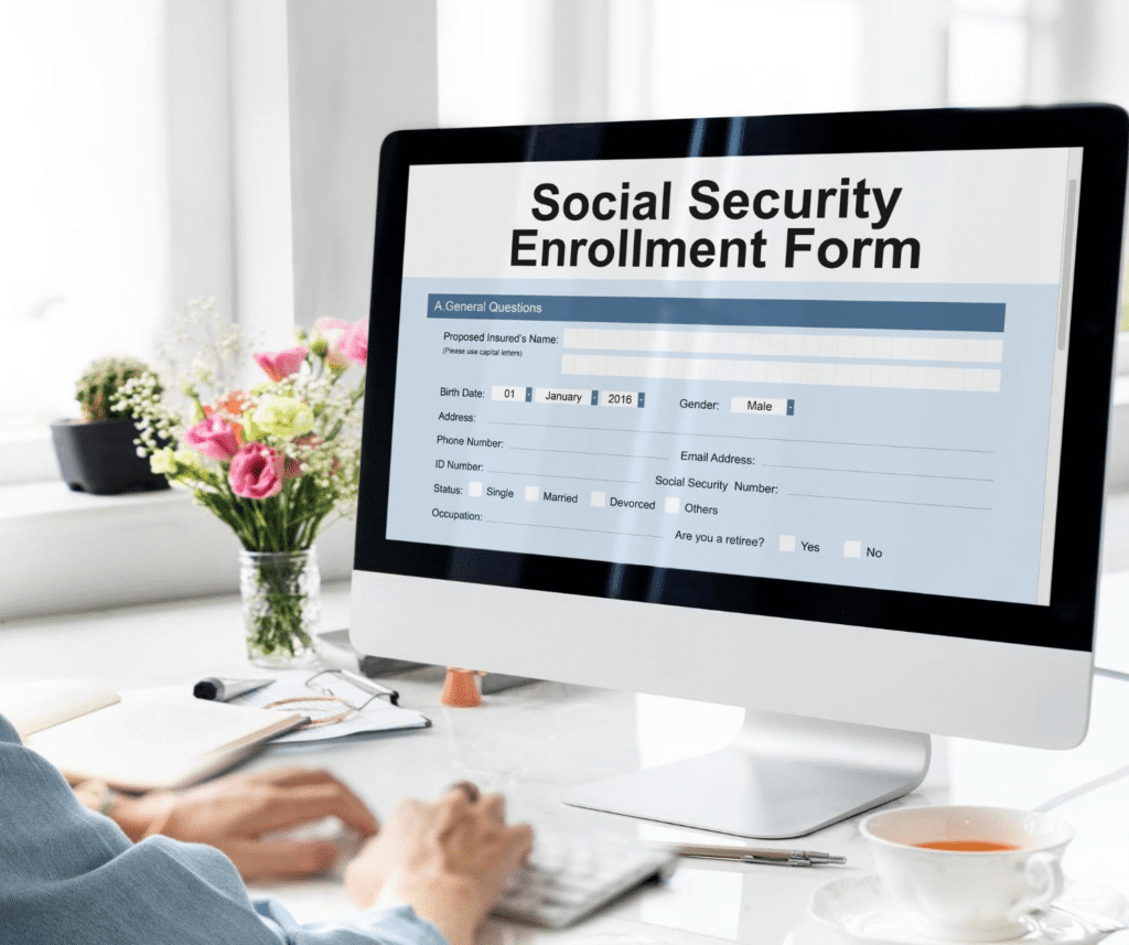 A person is typing on a computer in front of a monitor that displays a "Social Security Enrollment Form."