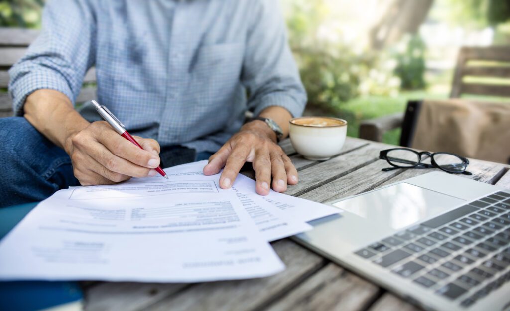 A man signs a list of forms placed on a garden desk along with his spectacles, coffee, and laptop