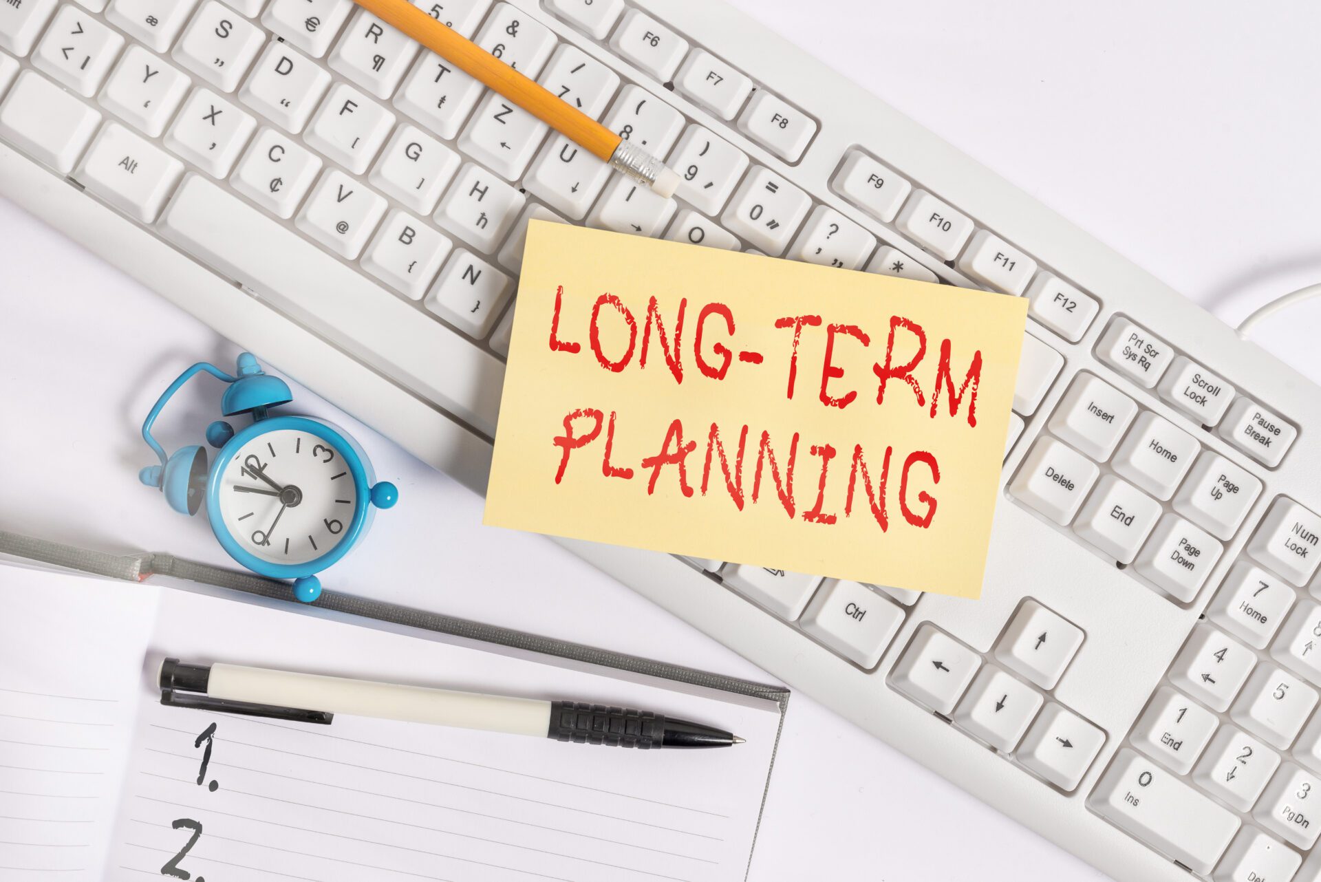 A text sign showing “long-term planning” is placed on a keyboard with a notepad, a clock, and a pen on the table.